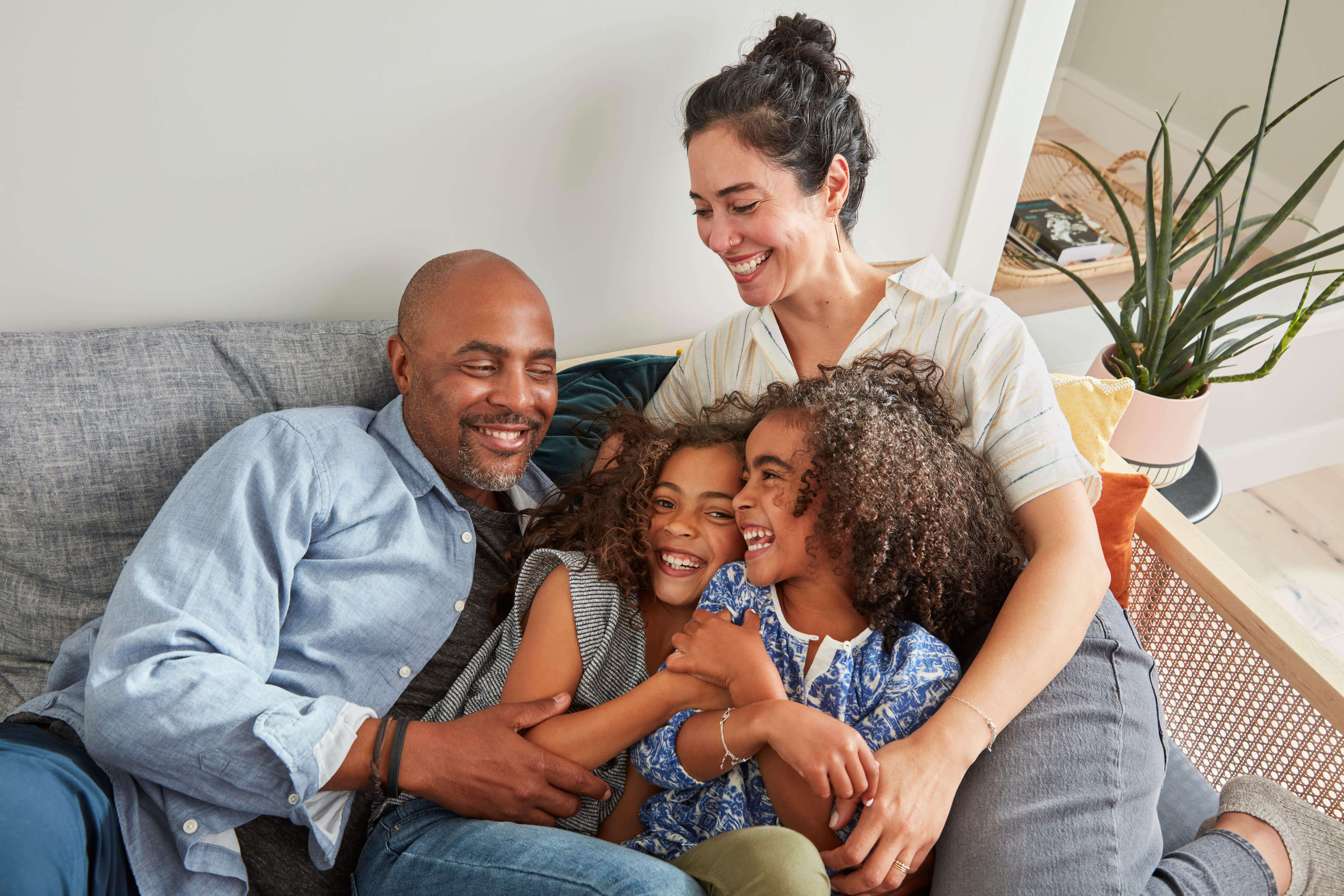 A happy group of four is sitting on a sofa and embracing. The man, wearing a light blue shirt, and the woman, in a striped shirt, are smiling at two girls, who are laughing and hugging each other. A potted plant is visible in the background, symbolizing growth and unity like California's CFRA.