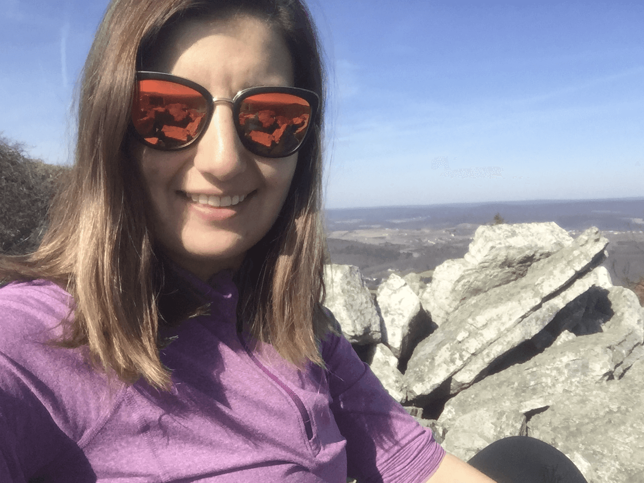 A person (Meaghan) with long brown hair wearing reflective sunglasses and a purple top is smiling at the camera. They are sitting outdoors on rocky terrain with a clear view of a distant landscape under a blue sky.