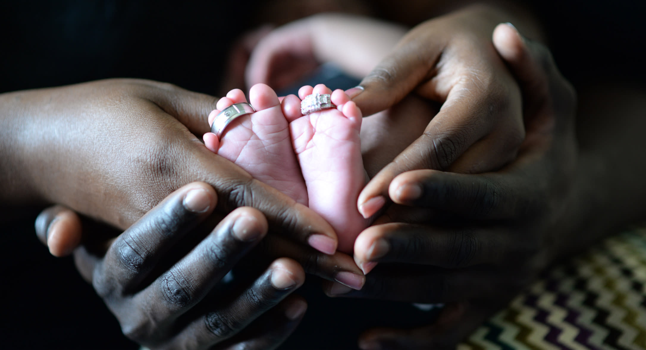 Close-up of two pairs of adult hands gently holding a baby's feet, each toe adorned with a ring. The image focuses on the contrast between the baby's soft skin and the adults' hands, emitting a sense of care and tenderness, reminiscent of the attention required during family moments like those covered under Family and Medical Leave Act.