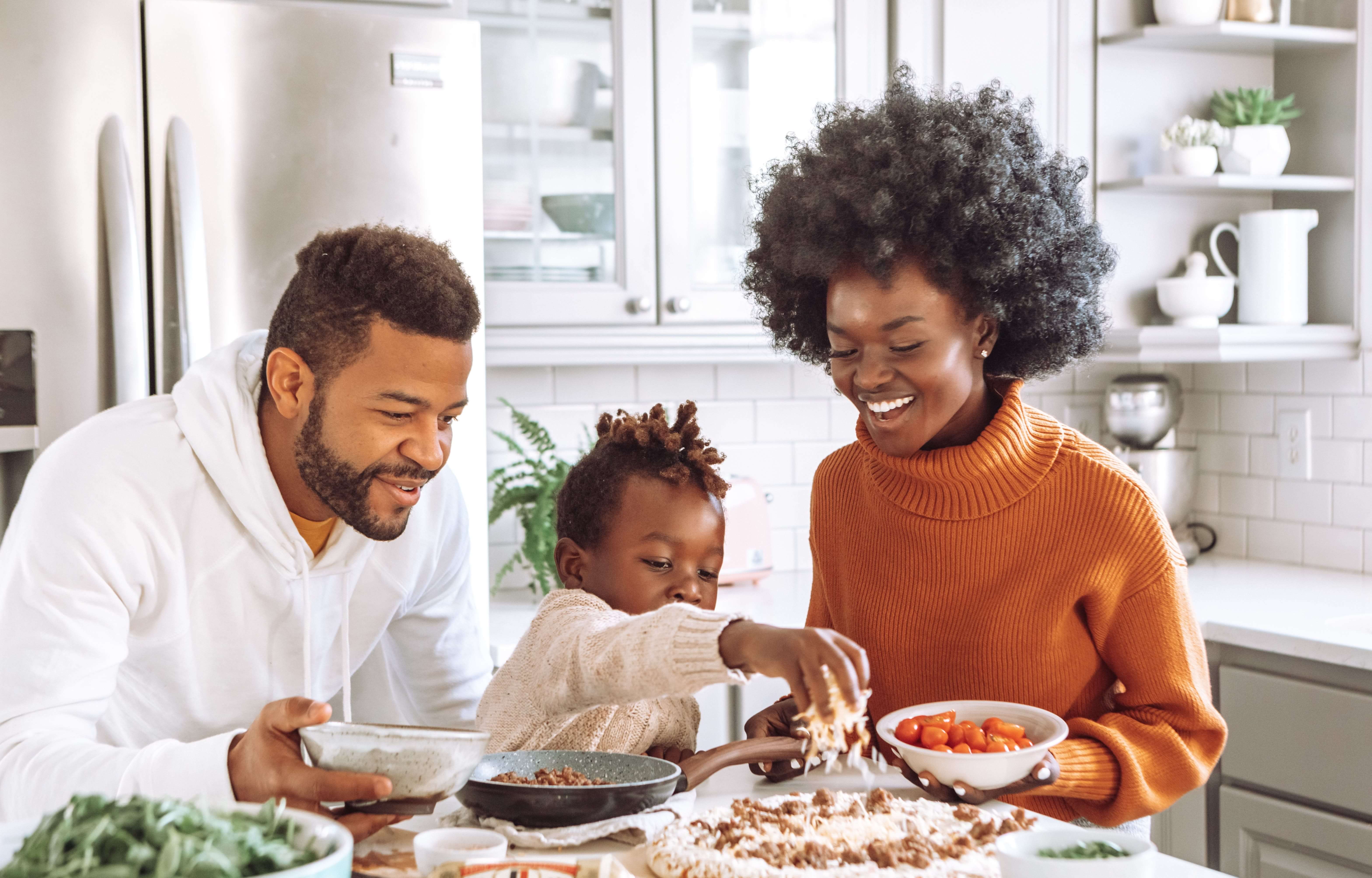 A family of three in a modern kitchen enjoys preparing food together. The child sprinkles ingredients from a bowl, while the mother and father smile and assist, celebrating the FMLA 30th anniversary. The kitchen has white cabinets, stainless steel appliances, and various ingredients on the counter.