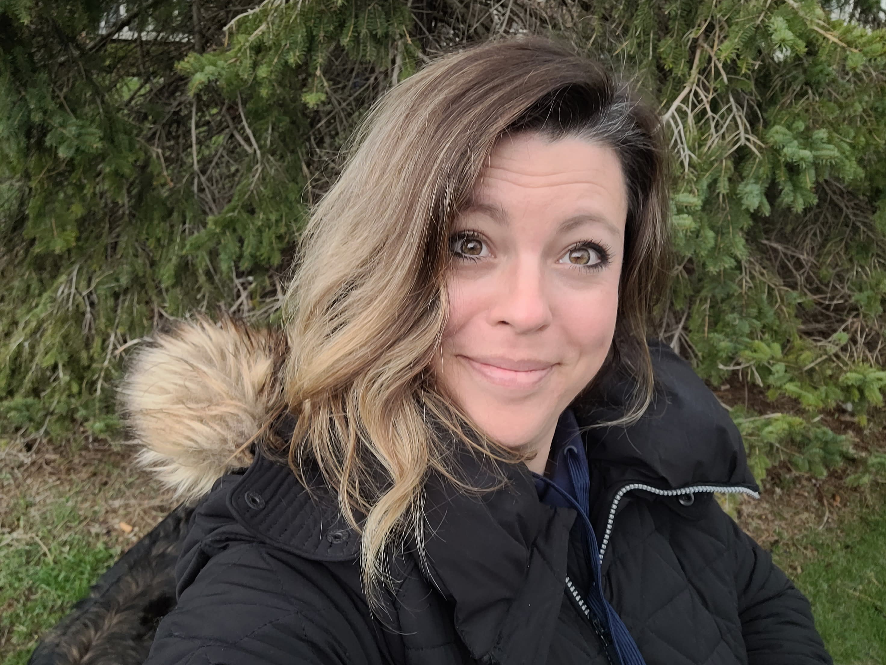 Katie, with her wavy, shoulder-length hair, smiles warmly at the camera. She is wearing a dark winter coat with a fur-lined hood. The background features green pine tree branches.