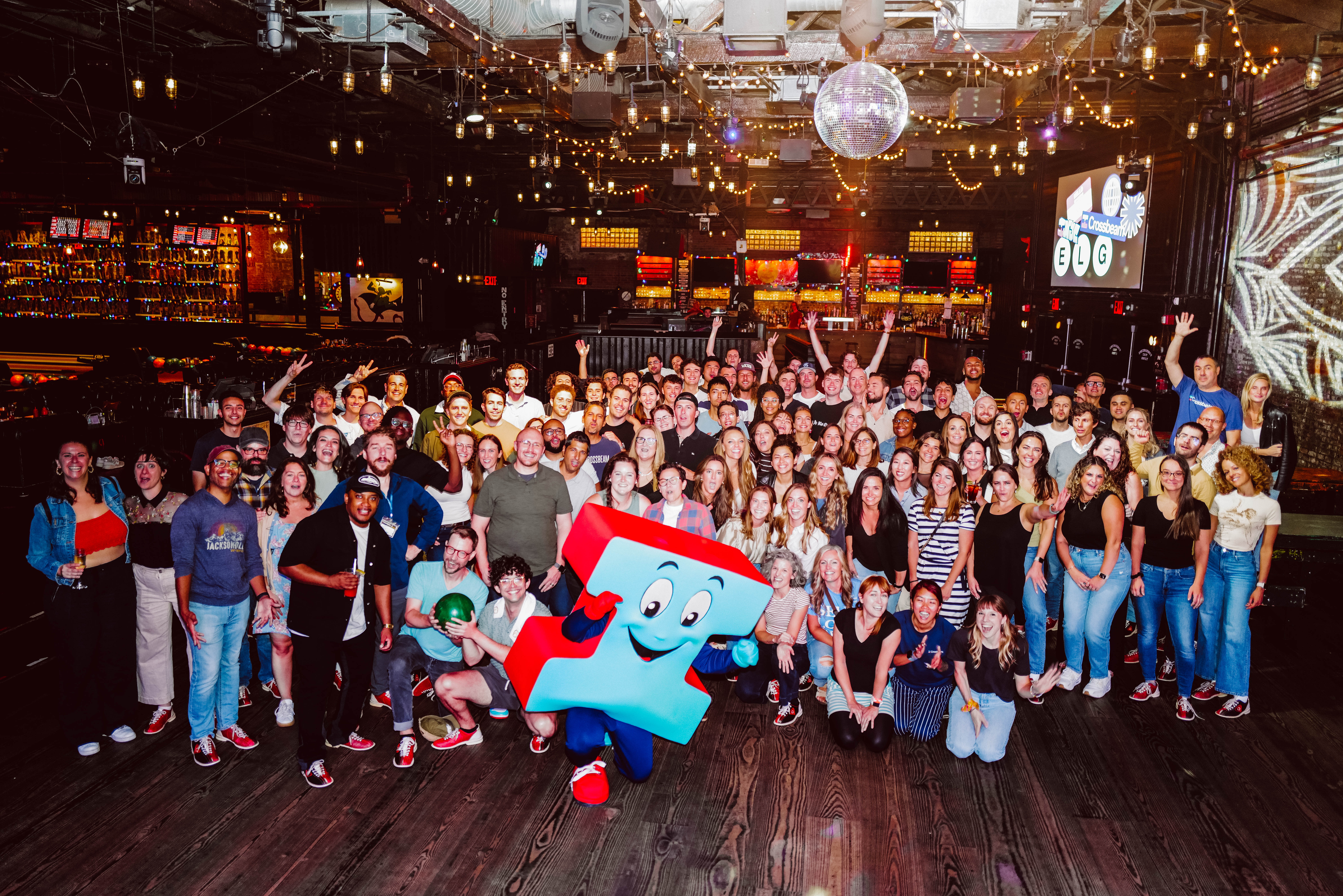 A large group of people pose together indoors, under festive lighting and decor that celebrates employee well-being benefits. Some hold drinks, with a person in a colorful mascot costume front and center. A bar and stage with equipment are visible in the background, adding to the joyful atmosphere.