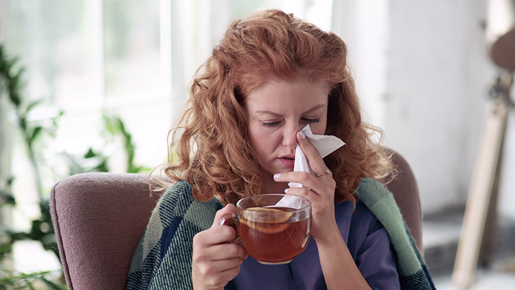 Person with curly red hair holding tea and tissue, looking unwell with a cold in a bright room with plants.