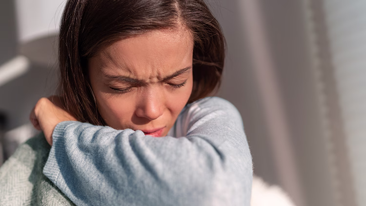 Person with furrowed brow and closed eyes in distress, wearing light blue sweater in warm sunlight.