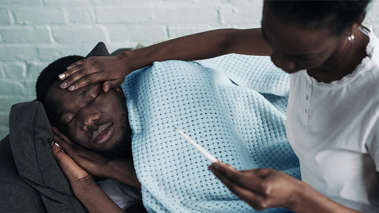 Close-up of a person checking temperature of sick individual lying on couch covered with blue blanket against white brick wall.
