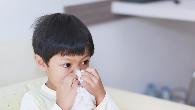 Young child with dark hair wiping nose with tissue, wearing white long-sleeve shirt in light-colored room.