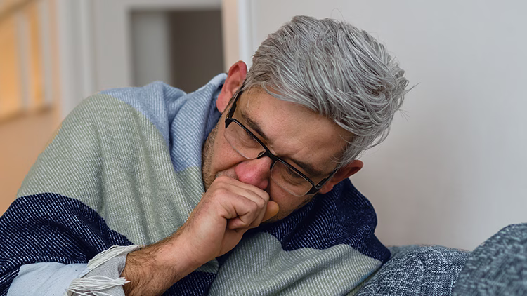 Person with gray hair and glasses coughing into hand, wearing a blue and green striped blanket or shawl.