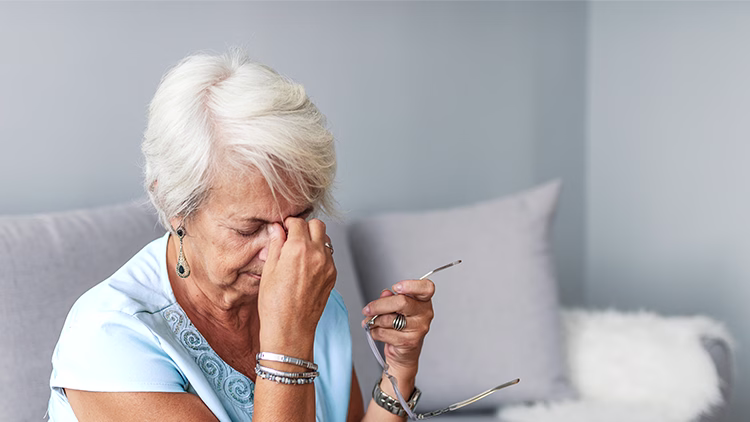 Gray-haired person in blue shirt holding glasses and pinching bridge of nose, showing signs of discomfort.