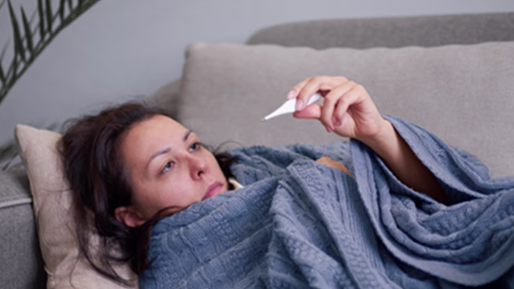 Person lying on couch wrapped in blue blanket, looking at thermometer in hand, appearing ill.