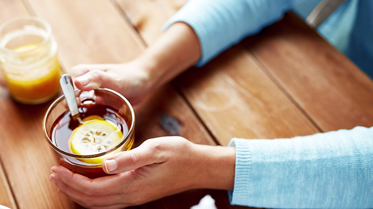 Hands in light blue sweater holding glass of tea with lemon slice, jar of honey nearby on wooden table.