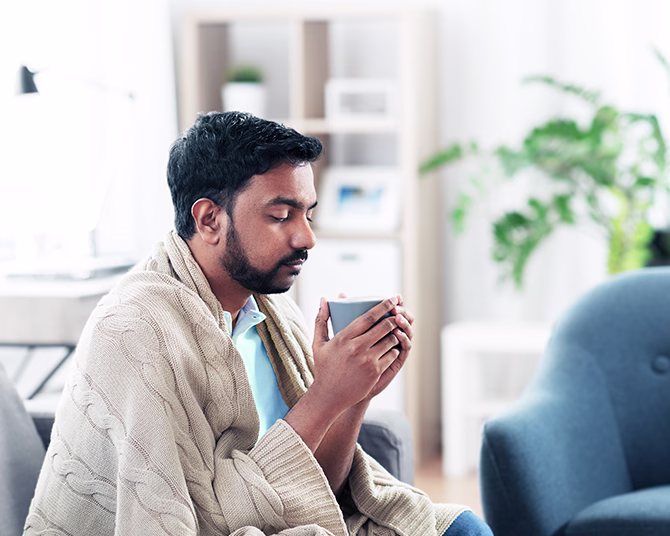 Person in beige sweater holding gray mug in bright living room with plants and bookshelves in background.