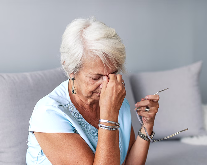 Gray-haired person in blue shirt holding glasses and pinching bridge of nose, showing signs of discomfort.