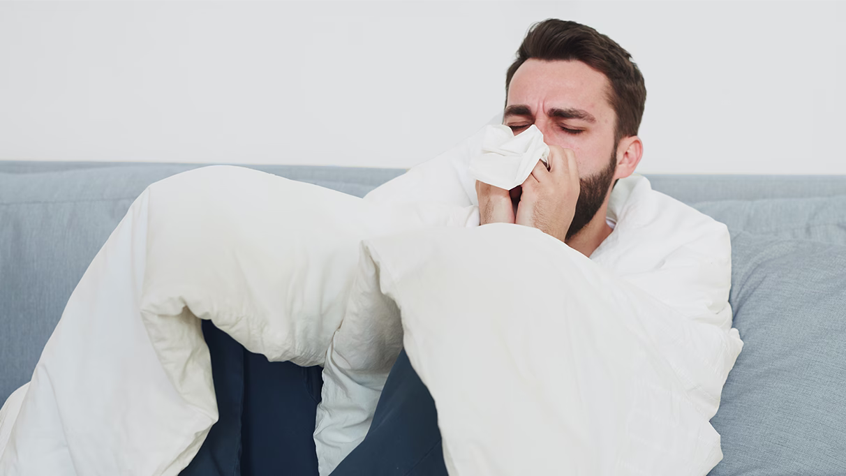 Person with beard blowing nose into tissue while sitting on light blue couch, wrapped in white blanket.