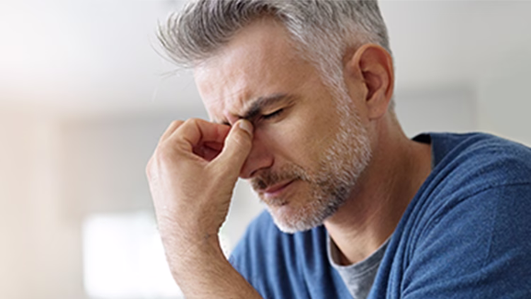 Close-up of a person with gray hair in blue shirt pinching bridge of nose, appearing stressed or experiencing discomfort.