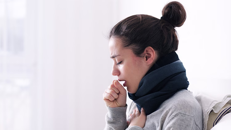Close-up of a person in gray sweater and blue scarf coughing with hand over mouth, appearing ill in bright room setting.