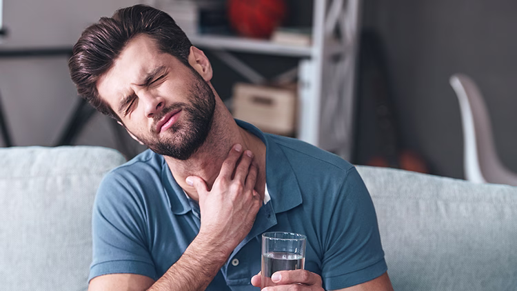 Person in blue shirt touching their neck in discomfort, appearing to have throat pain with a glass of water while sitting on a light-colored couch.