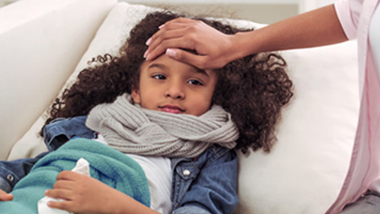 Close-up of child with curly hair lying in bed wearing scarf, covered with blanket as caregiver's hand checks forehead for fever.