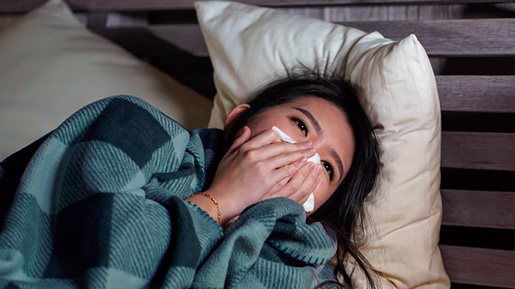 Close-up of a person lying in bed with blue blanket, holding tissue to nose, appearing sick with cold or flu symptoms.