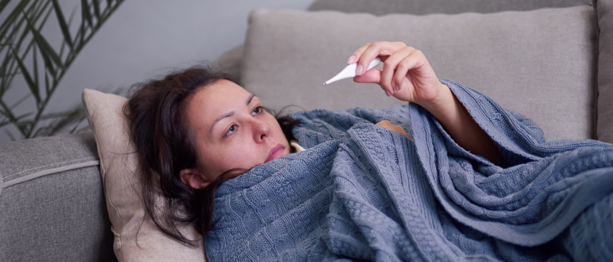 Person lying on couch wrapped in blue blanket, looking at thermometer in hand, appearing ill.