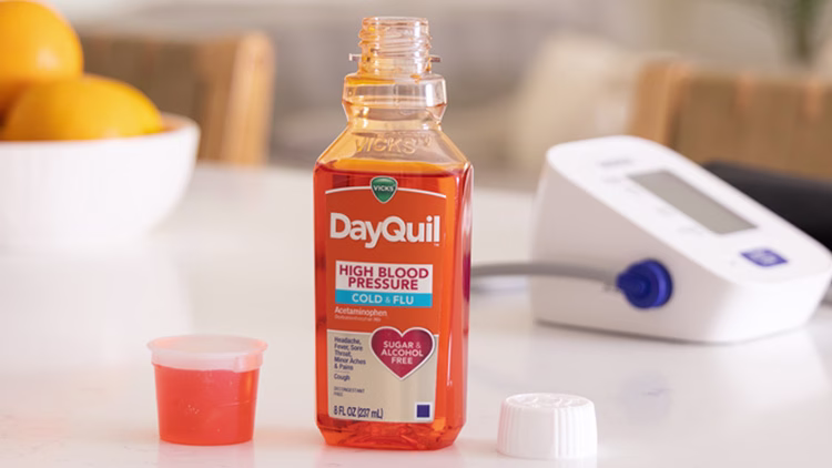 Open bottle of DayQuil High Blood Pressure Cold & Flu medicine with measuring cup, oranges in bowl, and thermometer nearby.