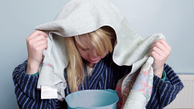 Person with towel over head leaning over blue bowl, wearing striped sweater against light blue wall.