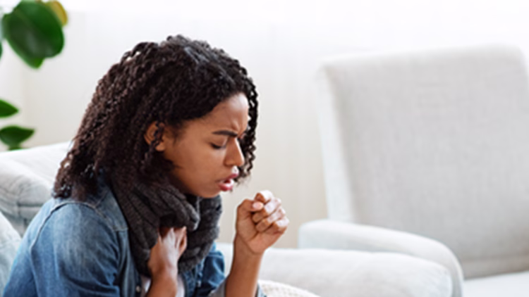 Close-up of a person with curly hair wearing denim and scarf coughing with hand on chest and coughing, appearing ill in bright home setting.