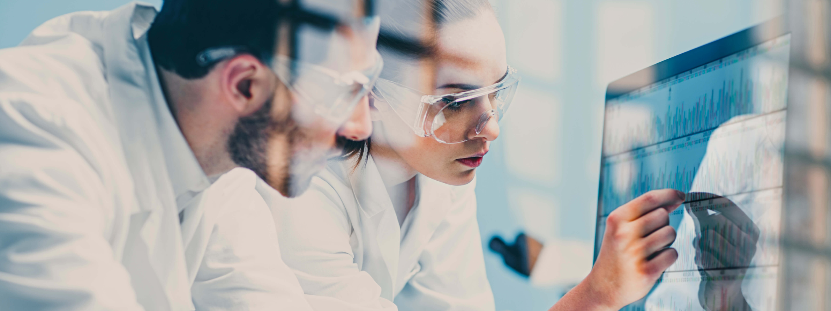 a stock photo showing a man and a woman wearing lab goggles in a lab type environment and pointing at some data in a screen