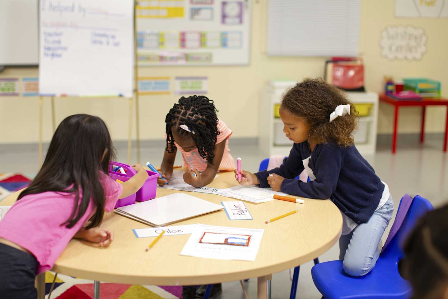 Kids drawing and scribbling around a desk in their classroom.