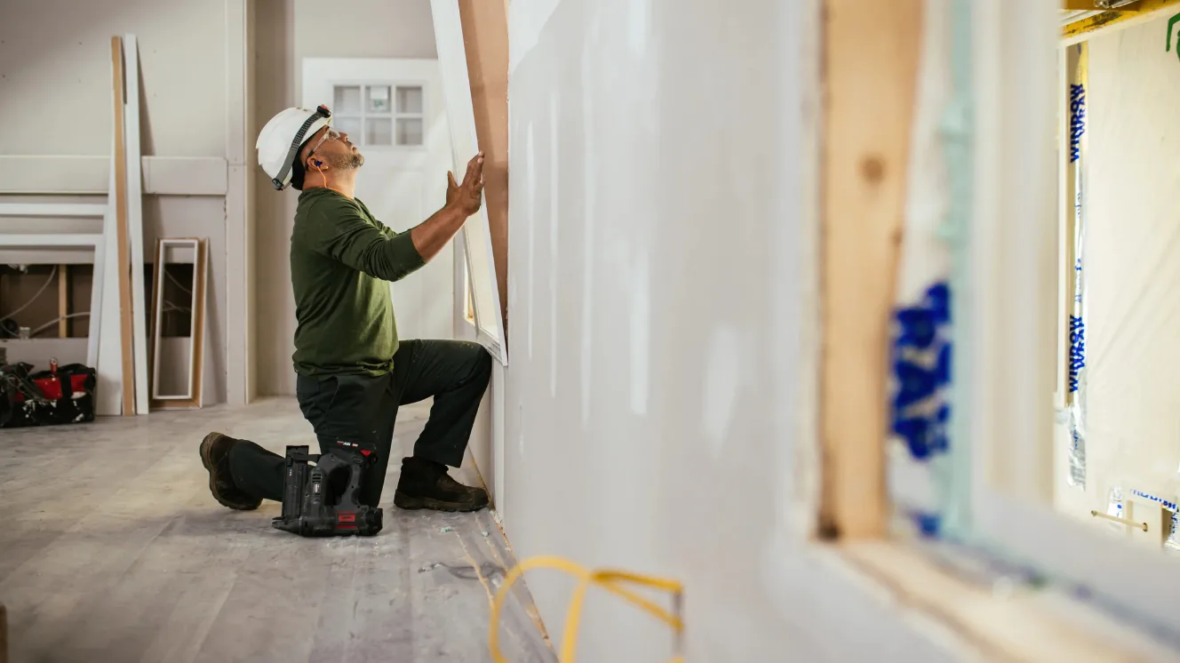 A man in a green shirt and construction hat kneels the floor of an under-construction home to hang drywall.