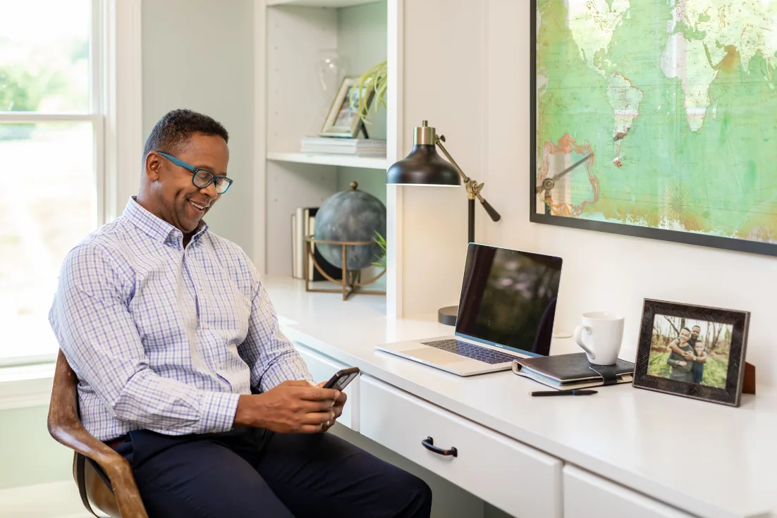 A smiling man in a checked button-up shirt and glasses sits at a white desk in a home office while looking down at his phone.
