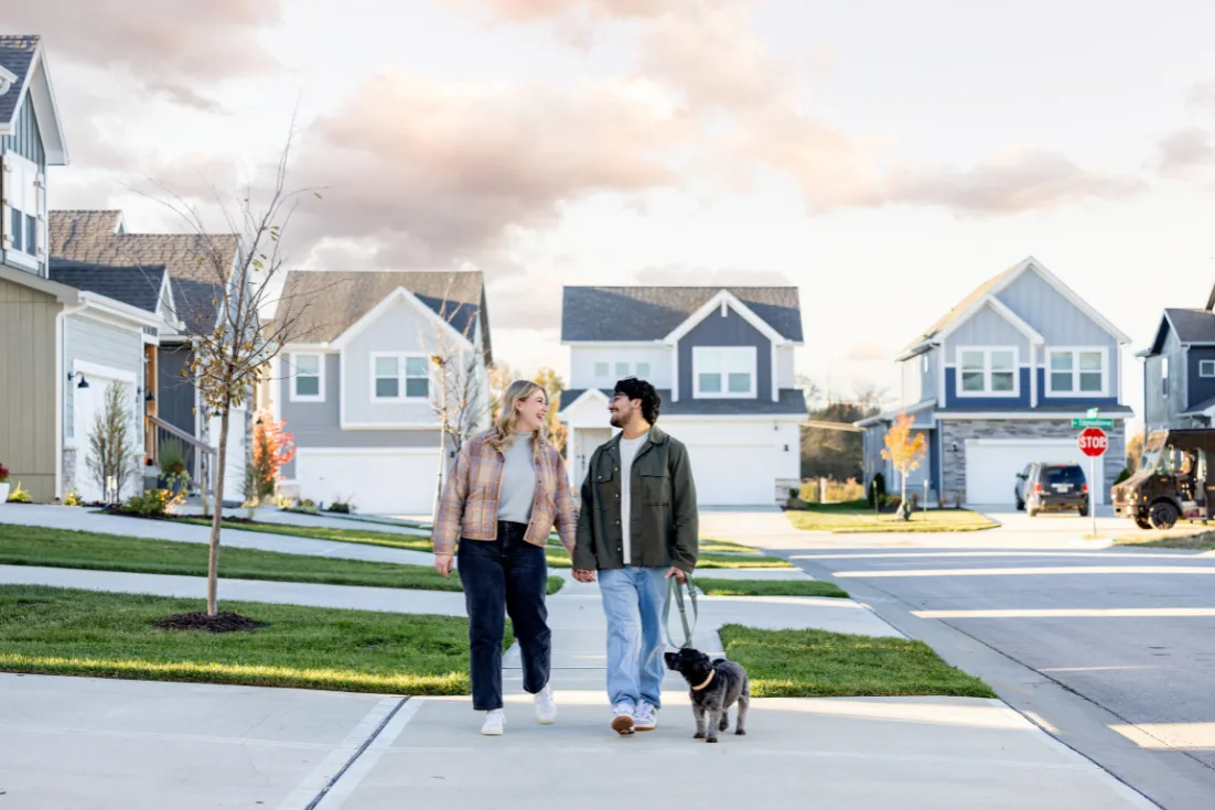 A couple in street clothes walks their dog on the sidewalk of a neighborhood with two-story homes behind them.