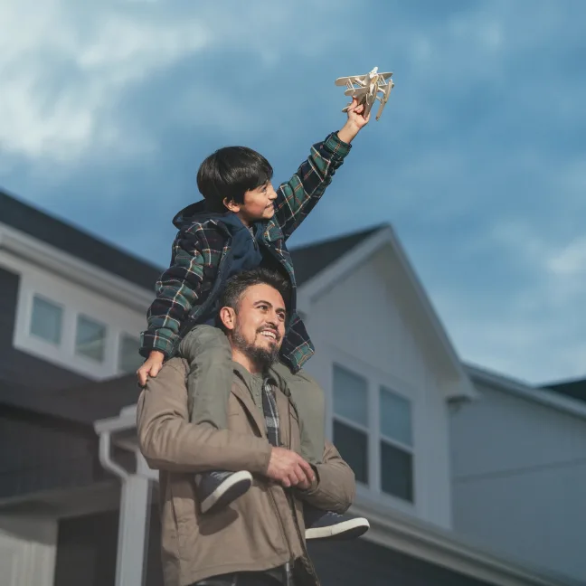 A boy sits on his dad's shoulders while holding up a toy airplane, a two-story home and a cloudy sky behind them.