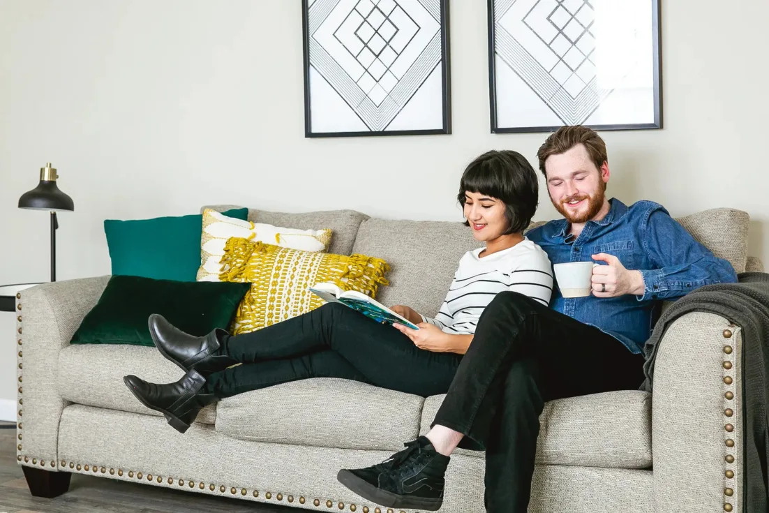 A couple relaxes on a beige couch with colorful throw pillows piled at the end.