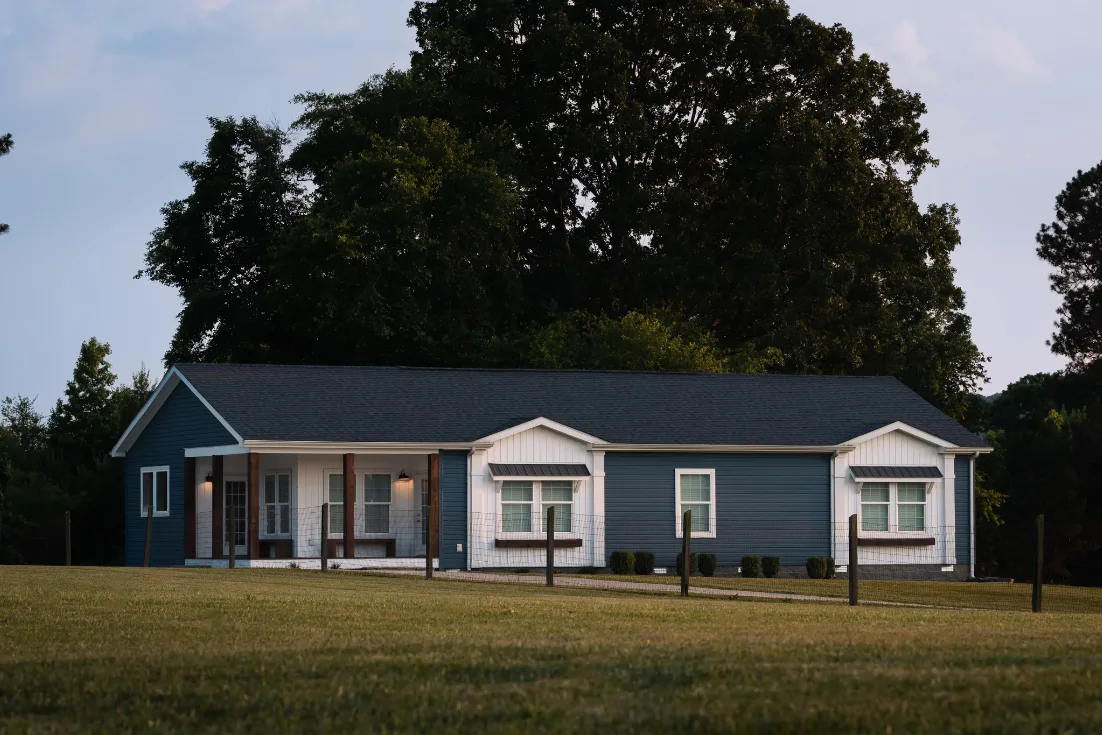 The exterior of a modern manufactured home with blue and white siding and side porch in the country at dusk.