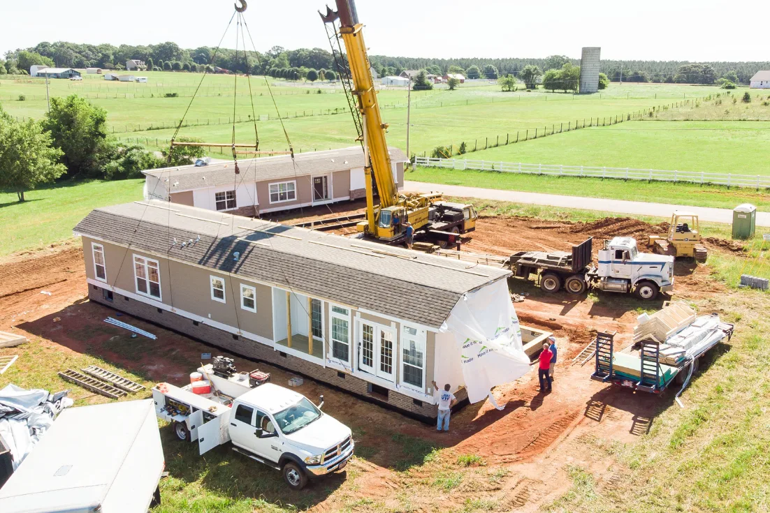 A manufactured home site with 2 home sections with gray-brown siding and white trim, crane, trucks and workers.