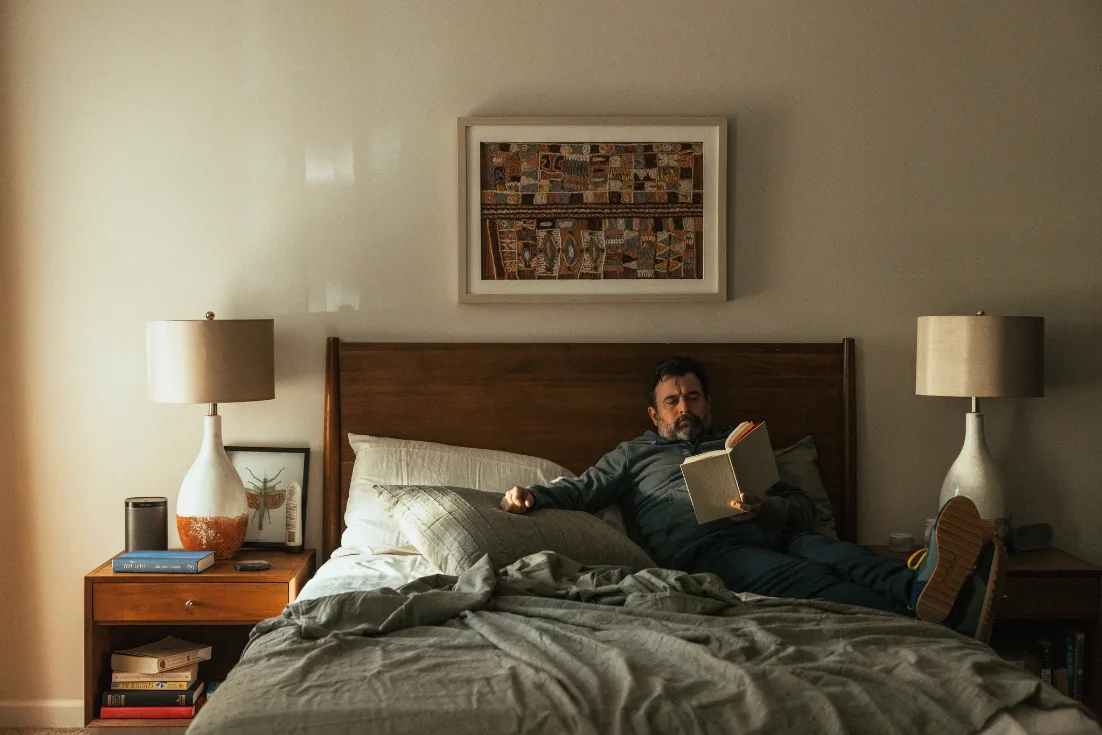 A man sits against the wooden headboard of his bed with his feet crossed while reading a book.