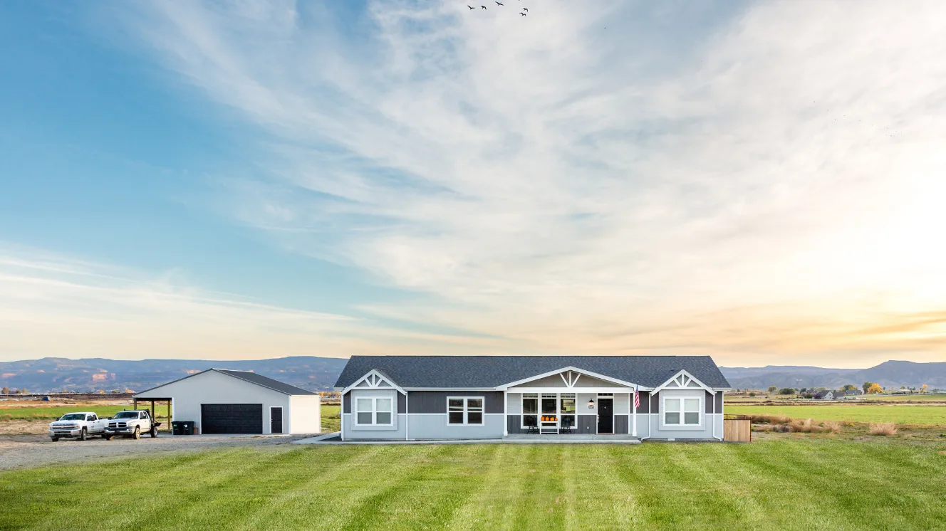 An exterior of a modern manufactured home with blue siding and a large front porch on a green field, a standalone work shed and two white trucks parked to its left.