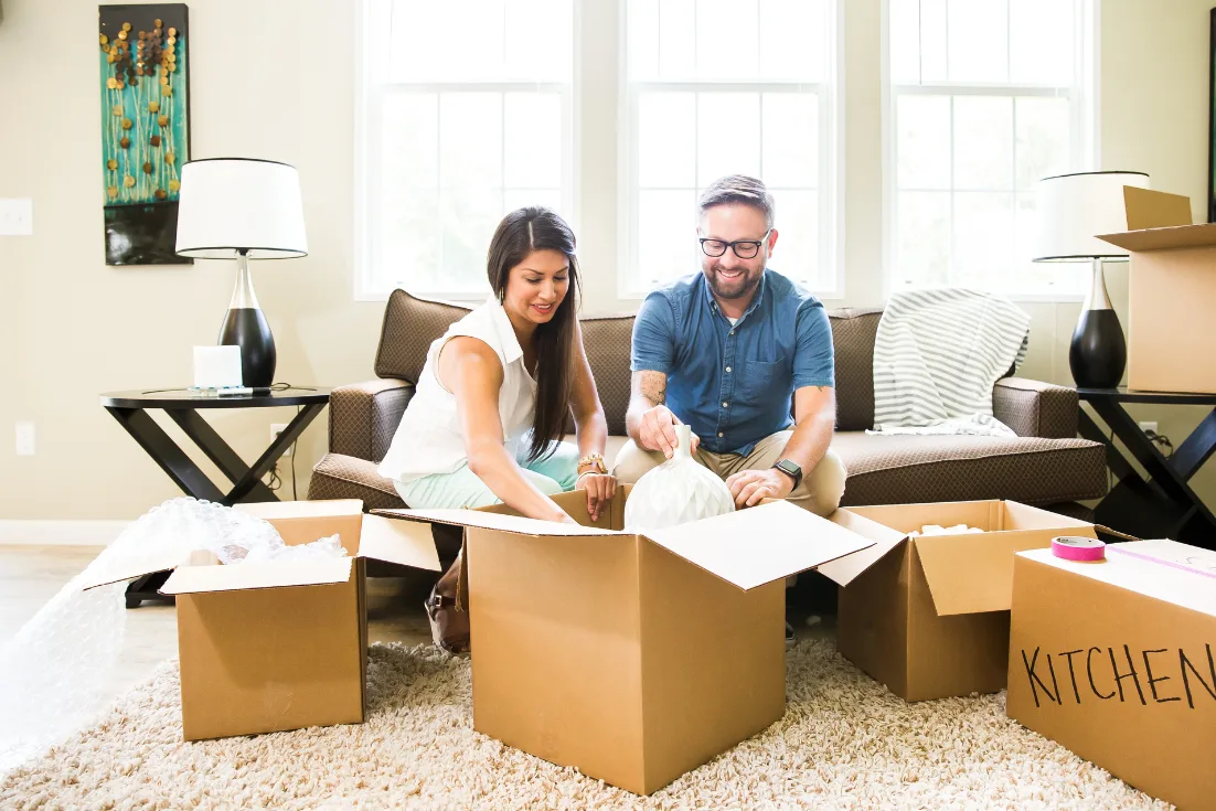  A couple sits on a leather couch placing items in one of the many moving boxes in front of them.
