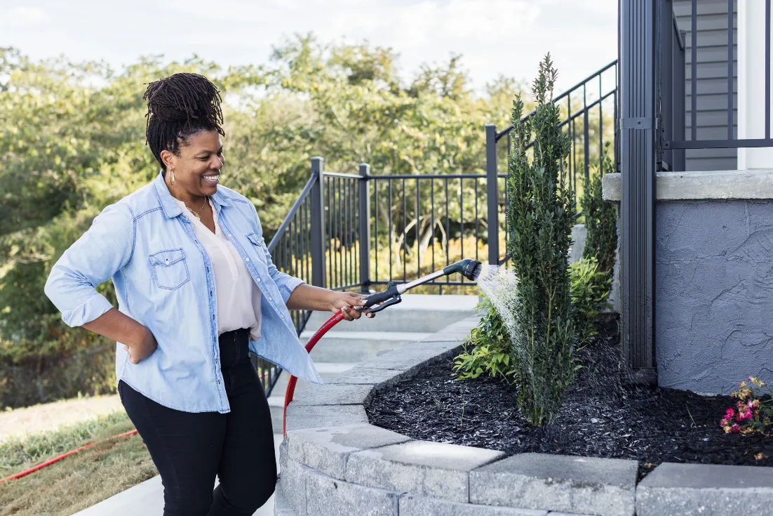 A woman in a chambray shirt smiles while watering her flower bed with a hose.