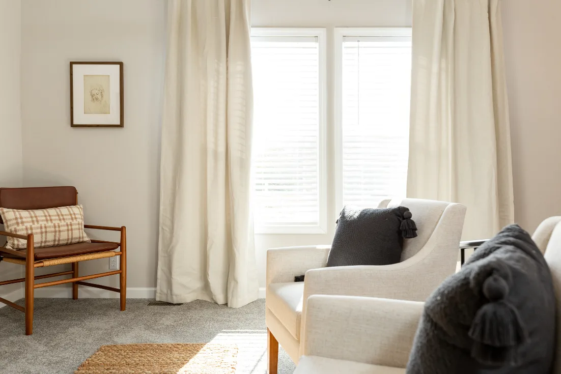 A living room features a modern leather chair in one corner, tall double windows hung with curtains, and white upholstered arm chairs.