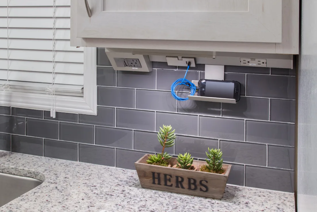 A close-up of a charging station under a white cabinet and against the gray subway-tile backsplash of a kitchen that hangs above a square potted plant painted with the word "herbs."