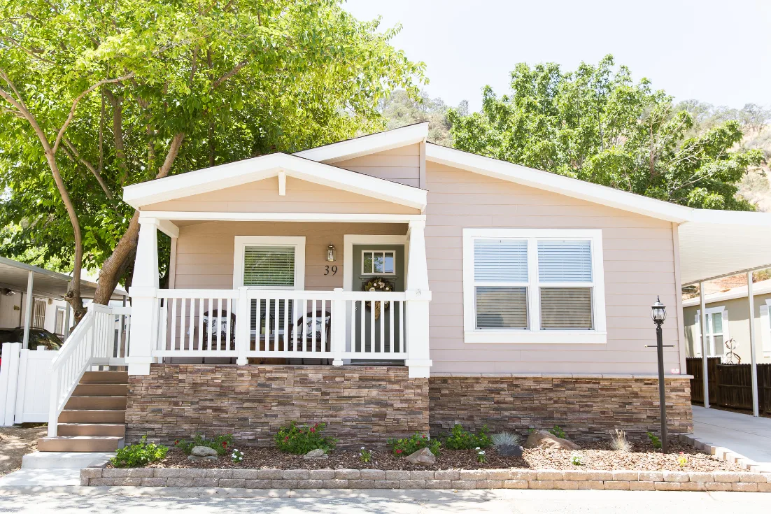 An exterior of a Craftsman-style manufactured home with beige siding, brick skirting, and a front porch with white railing.
