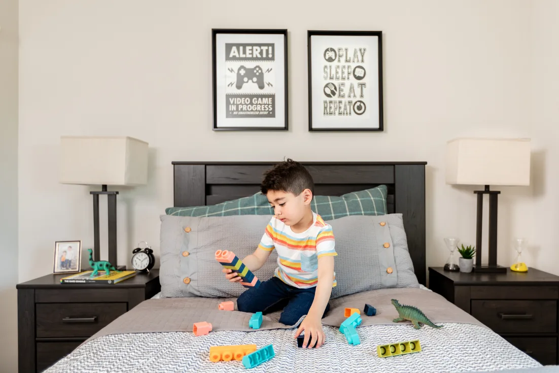 A small boy in a colorfully striped shirt sits on his bed playing with blocks, while more blocks and a toy dinosaur surround him on the gray comforter.