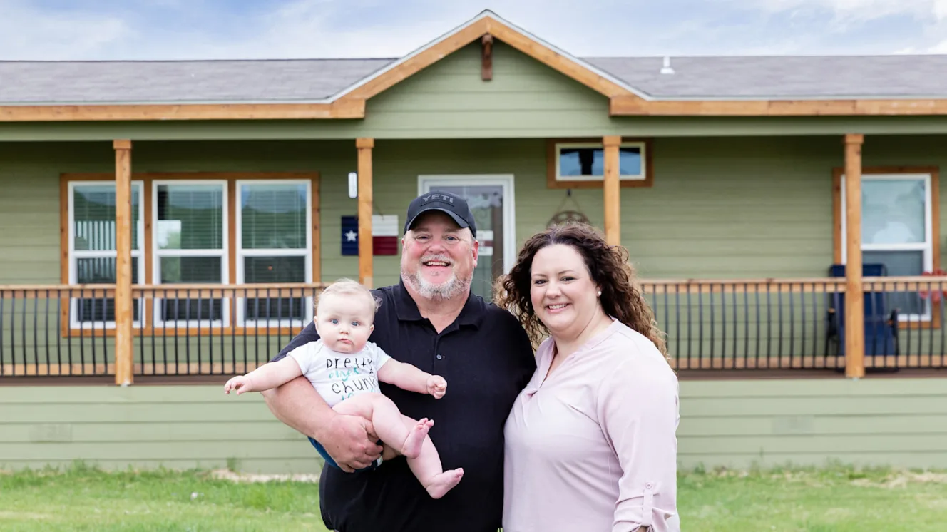 Couple stand in font of their forest green cabin home with wide front porch with man holding their new baby.