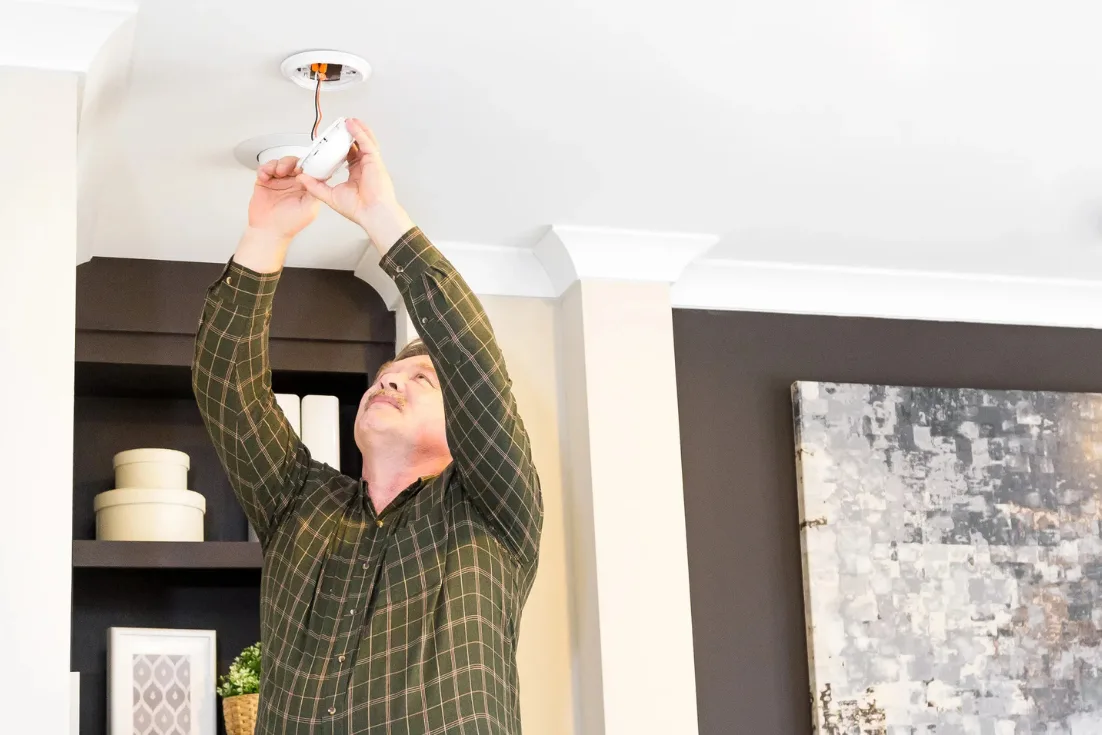 A man in a plaid shirt is reaching up to the ceiling and checking the smoke detector.