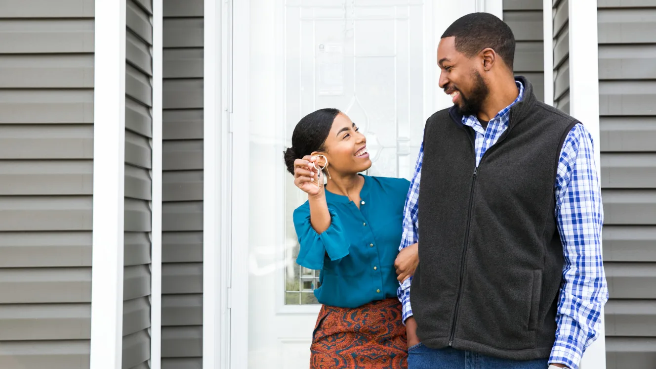 A woman in a blue blouse and a man in a plaid shirt and vest stand in front of a white door with their arms linked and turned to each other smiling while the woman holds up a home key.
