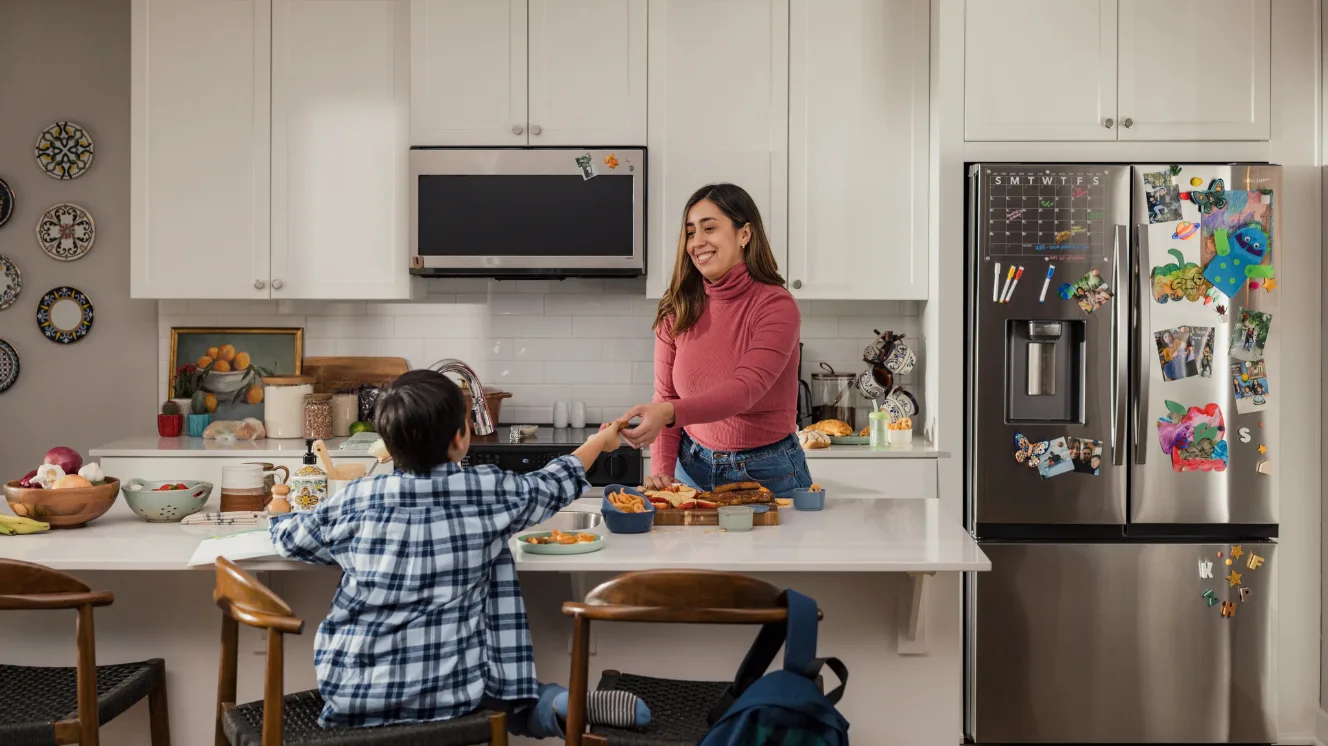 In a kitchen with white counters and cabinets, a mom in a pink sweater hands a snack to her son seated at the island to the left of a stainless steel fridge decorated with magnets, photos, and artwork.