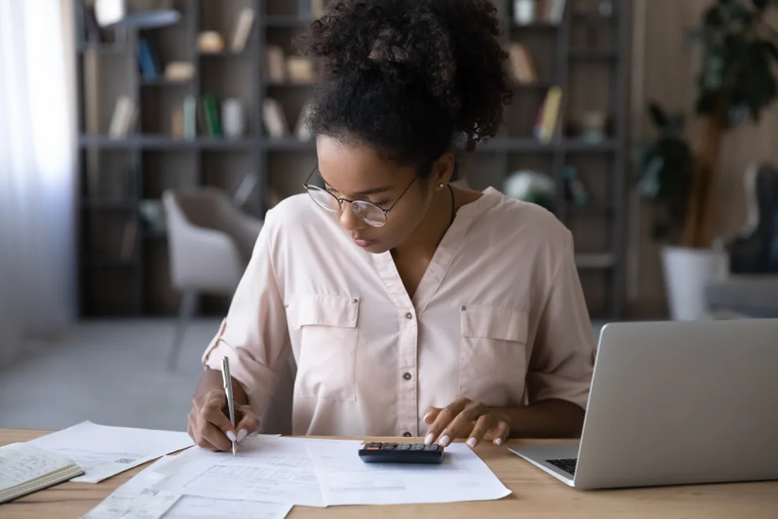 A woman in a pink blouse and glasses sits at a desk writing on papers with her right hand while tapping a calculator with her left hand beside a laptop.
