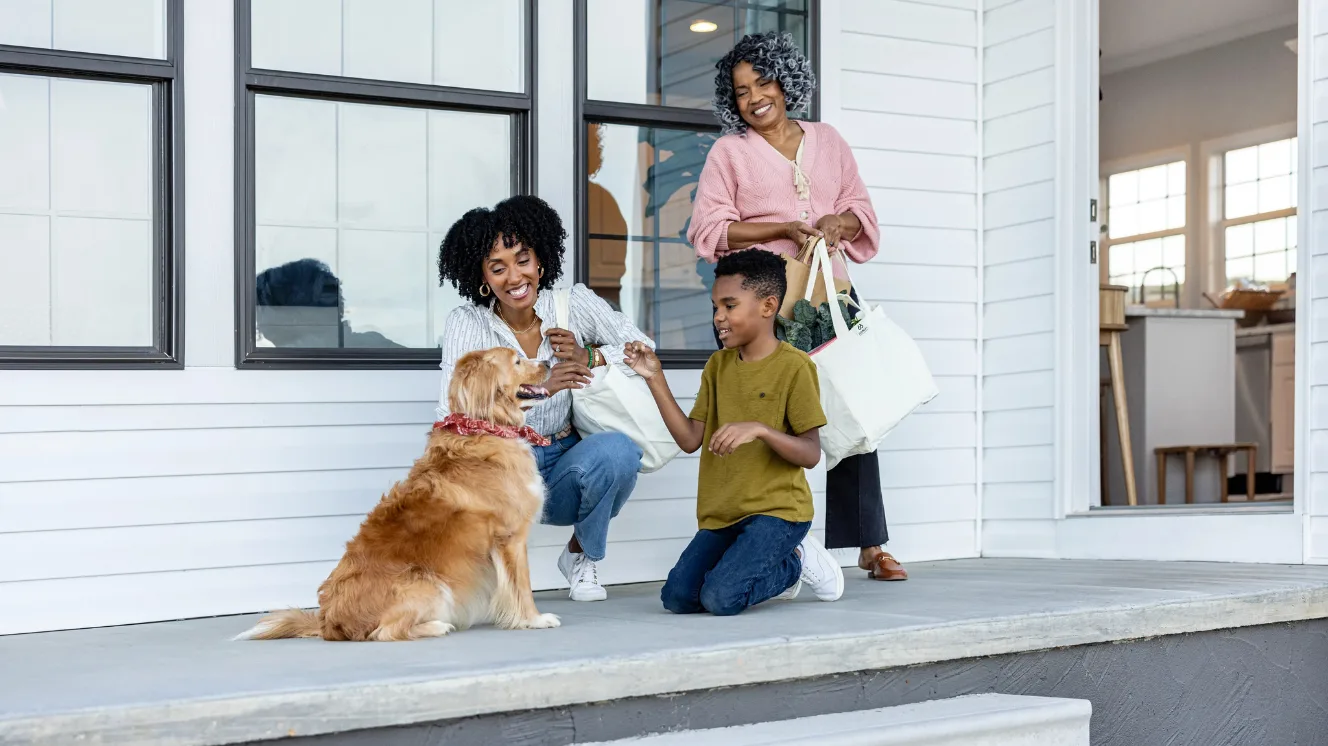 On the front porch, a dog sits for a boy kneeling and holding a treat while his mom, crouching down, and his grandmother, standing behind watching, both watch and smile.