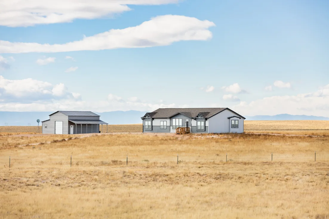 The exteriors of blue modern manufactured home and a detached shed shown on a grassy stretch of land under a blue sky.